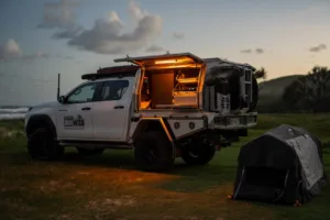 Toyota Hilux dual cab with Norweld canopy illuminated at a coastal campsite at dusk